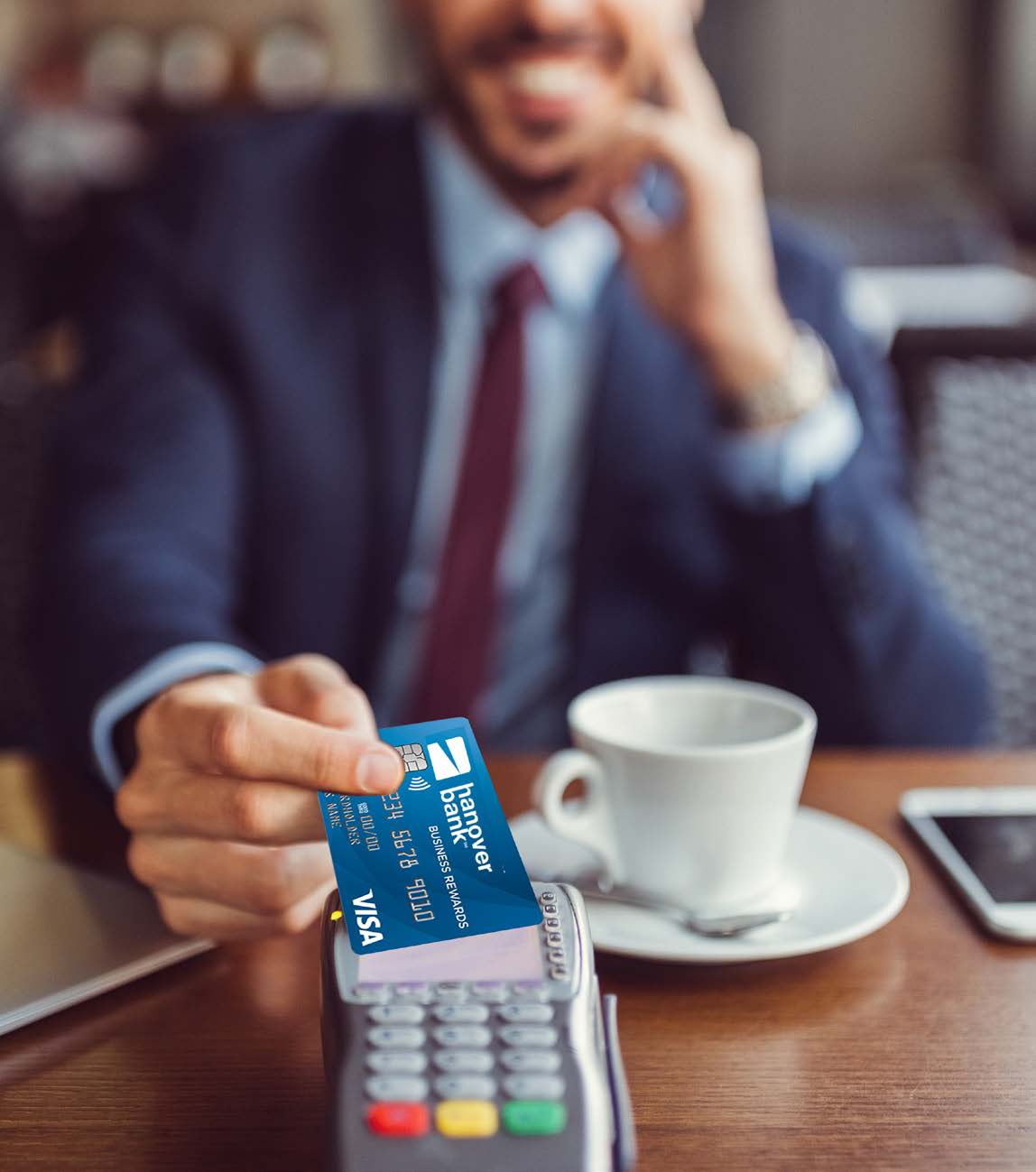 A person in a suit taps a blue Hanover Bank credit card on a payment terminal at a café table, with a cup of coffee and smartphone nearby; the mood is professional.
