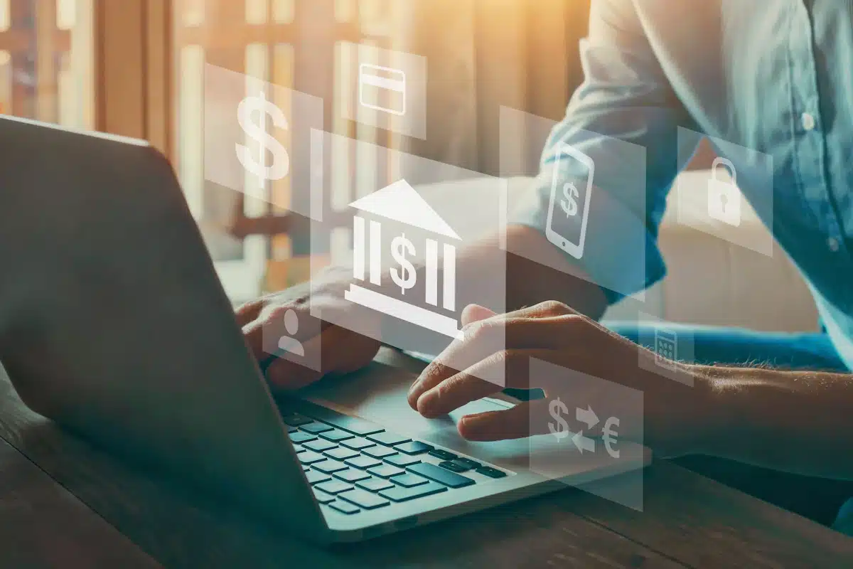 A person typing on a laptop with digital banking icons like a bank symbol, dollar signs, and mobile device overlaying the image, suggesting online banking.