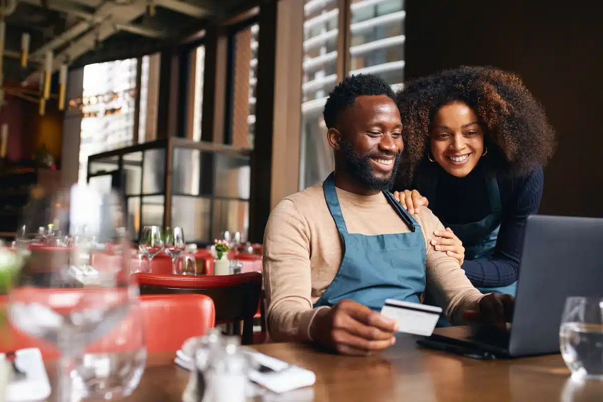 A cheerful man and woman wearing aprons smile as they use a laptop in a warm, cozy restaurant; the man holds a credit card while completing an online payment.
