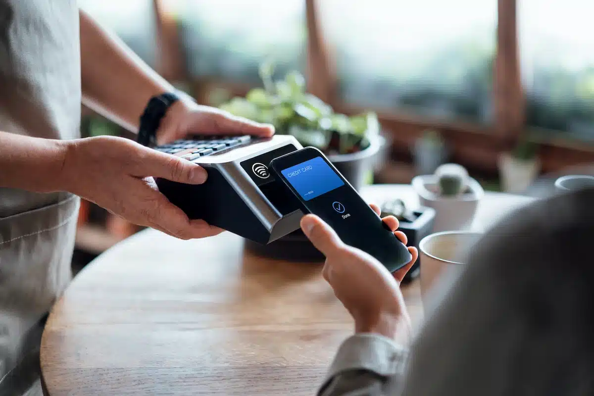 A person makes a contactless payment with a smartphone at a point-of-sale terminal, held by another person indoors. The setting is casual and modern.