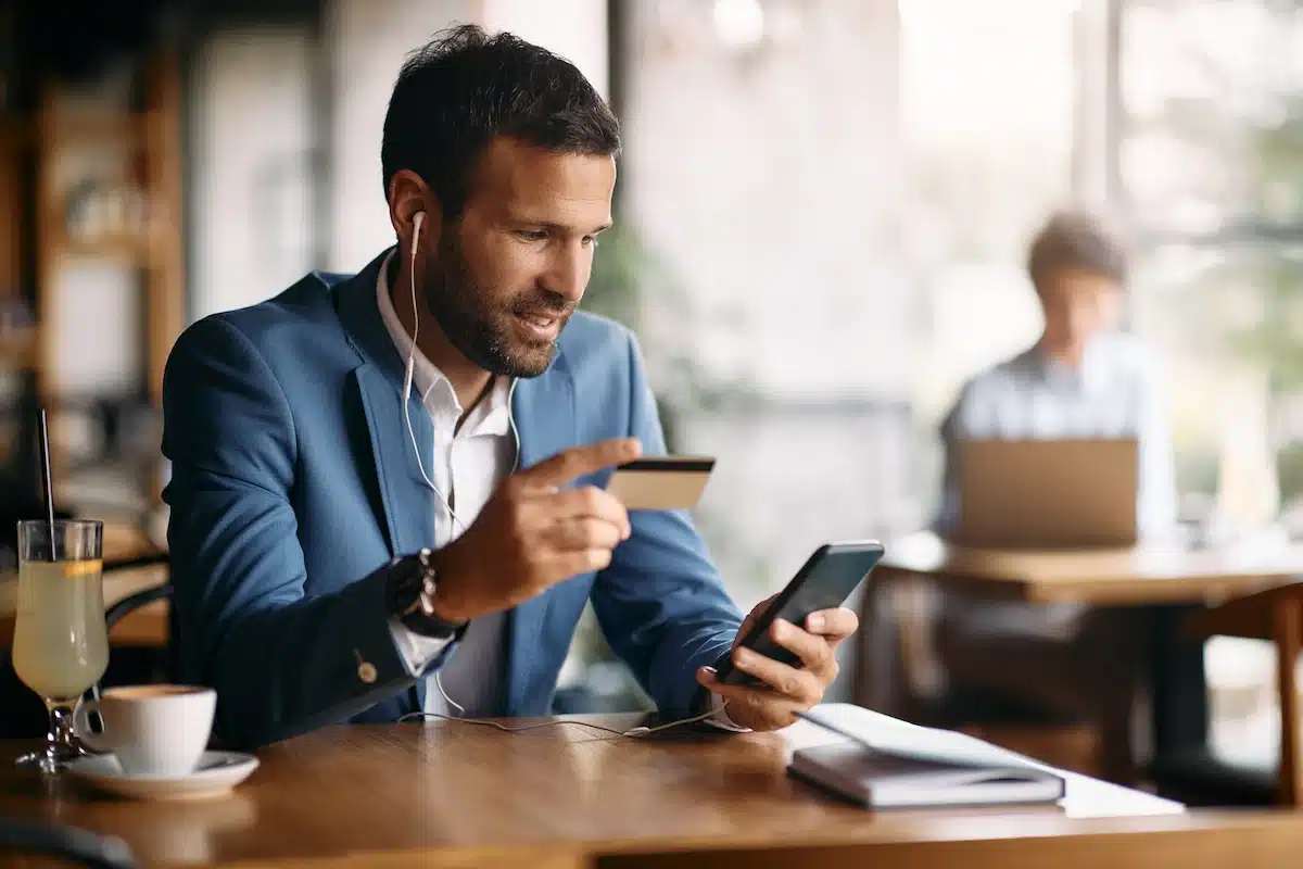 A man in a blue blazer sits, wearing earphones, at a cafe table, smiling and holding a credit card while looking at his phone. ,