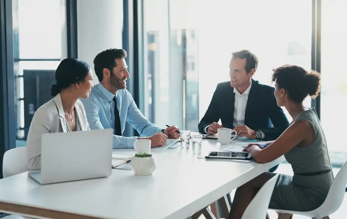A diverse group of four colleagues in a professional meeting sits around a bright conference table, engaged in discussion, with notebooks and laptops open.