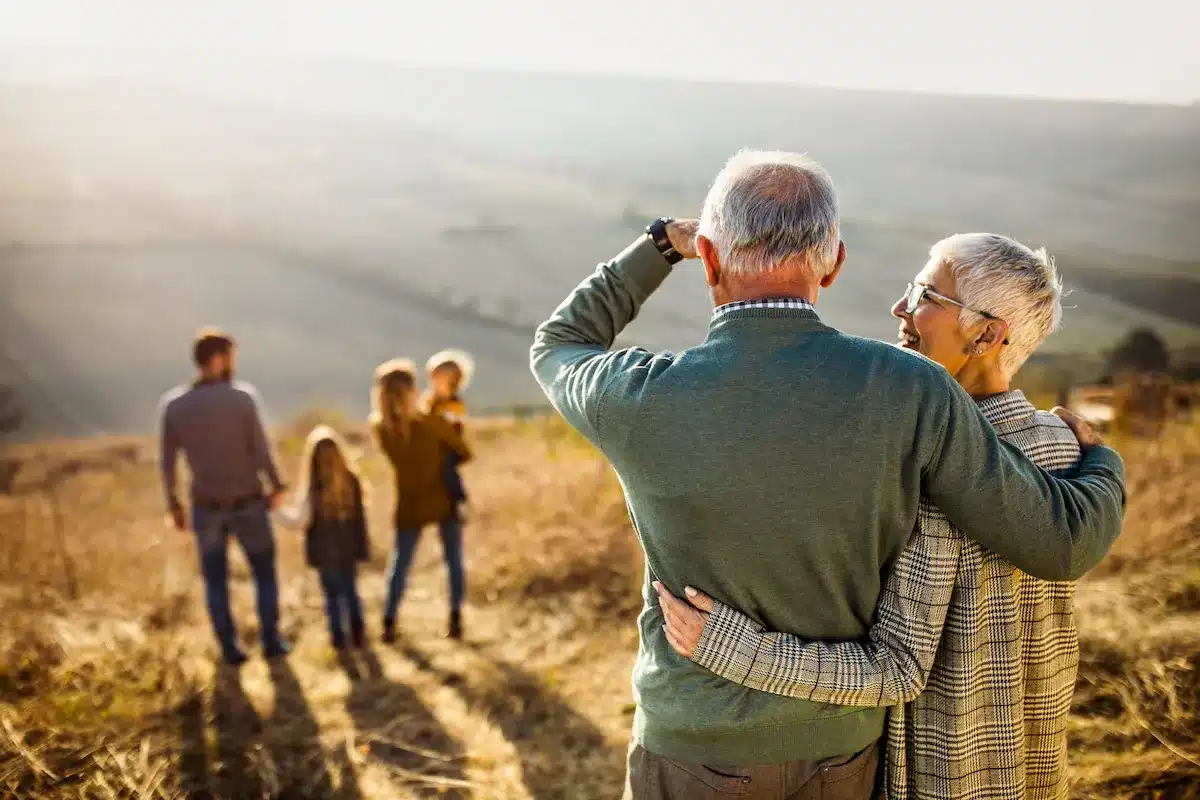 Elderly couple embraces and looks at a family with children in a sunlit field. Warm, peaceful atmosphere with golden light and rolling hills.