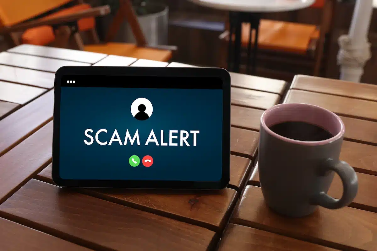 A tablet on a wooden table displays "Scam Alert" with call icons, evoking caution. A mug filled with coffee sits beside it, hinting at a casual setting.