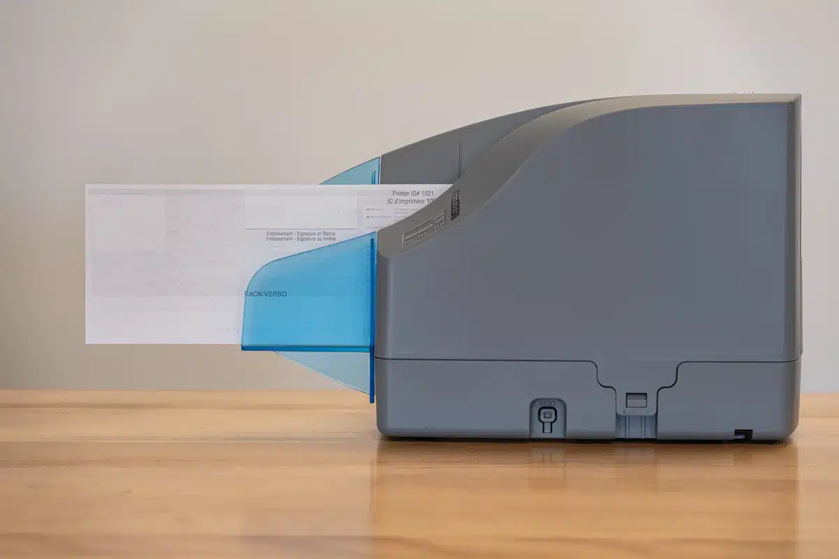 A grey check scanner sits on a wooden table. A check protrudes from the machine, partially visible in the blue semi-transparent output tray.