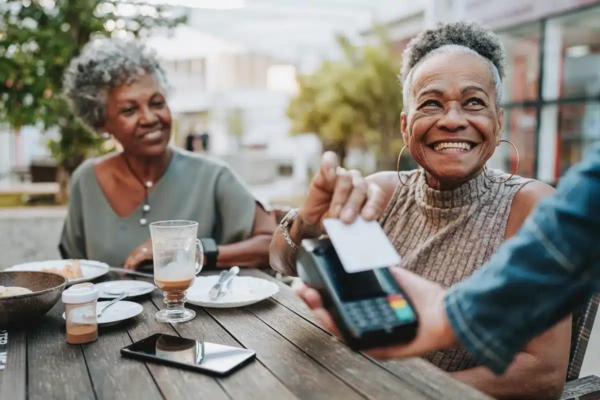 Two smiling women at an outdoor café table as one pays with a card reader. The atmosphere is joyful and relaxed, with beverages and tableware visible.