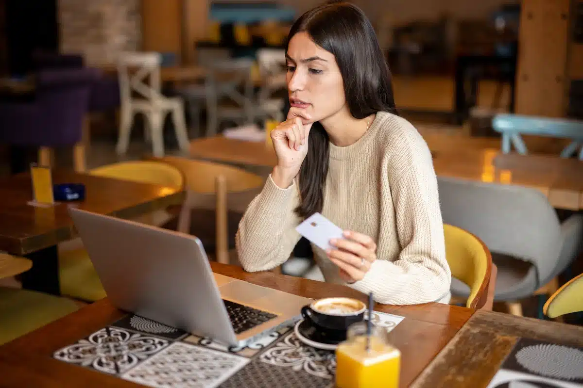 Woman in cafe focused on laptop, holding a credit card. Tables and chairs in background, with coffee and orange juice on wooden table.